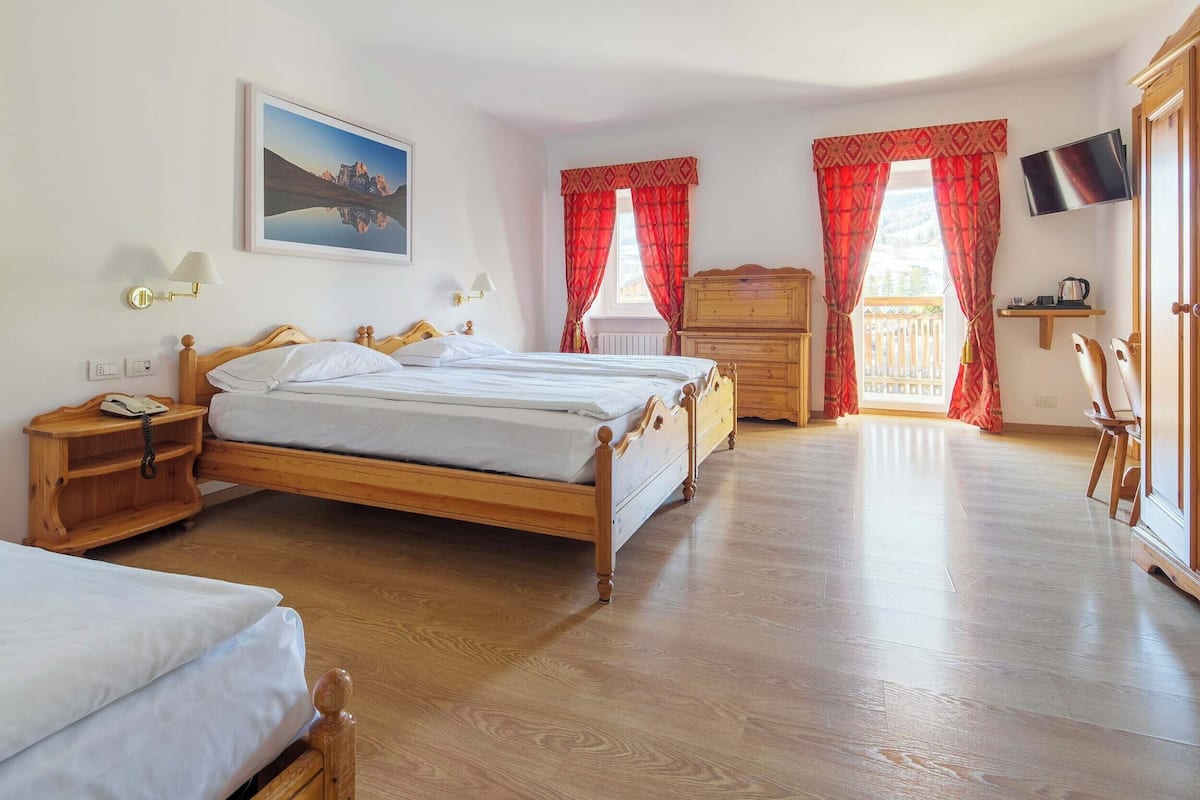 Un dormitorio con dos camas de madera, un aparador de madera, un televisor montado en la pared y una ventana con cortinas rojas.