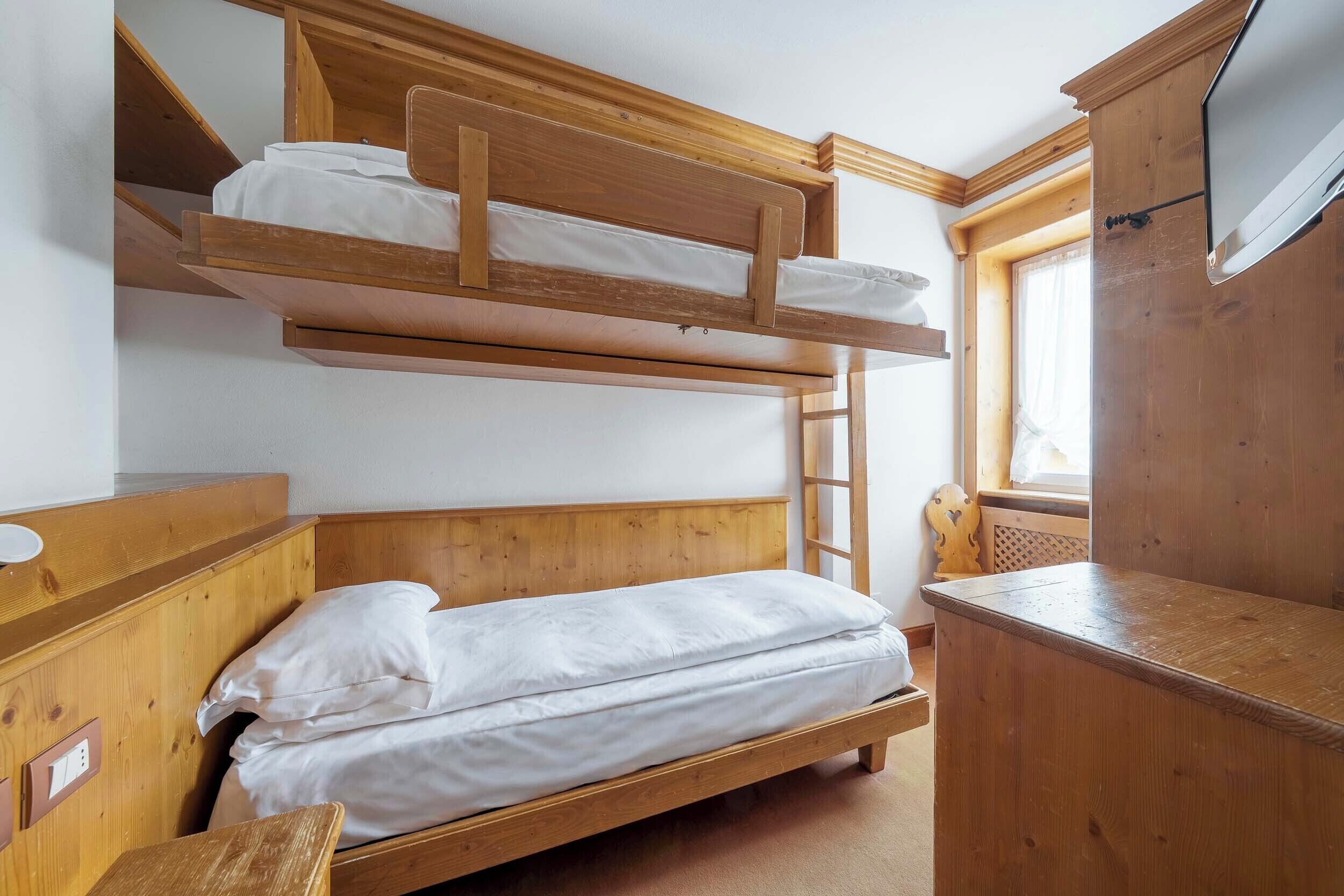 A bunk bed with a wooden headboard, a bedside table, a TV mounted on the wall, and a window with curtains.