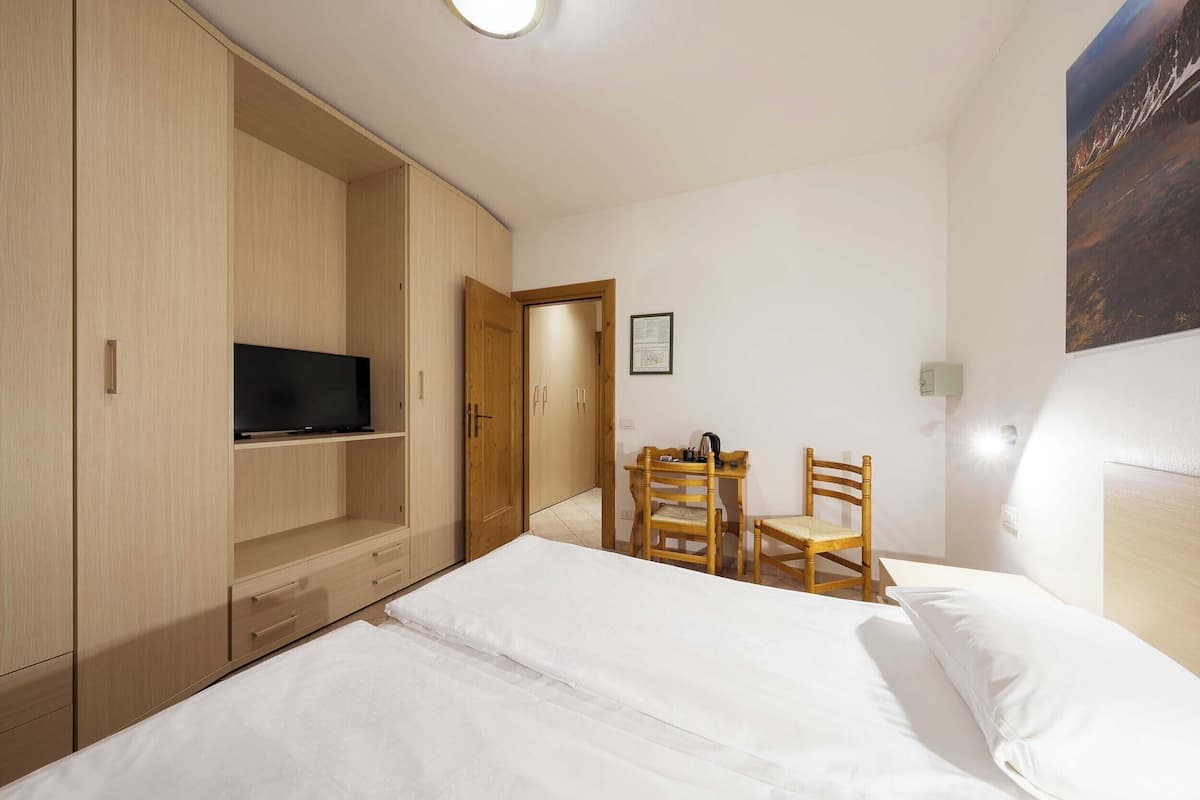 Un dormitorio con cama, televisor en estante, silla de madera y puerta que da a otra habitación.