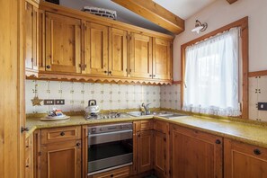 A kitchen with wooden cabinets, a built-in oven, a sink, and a window with a curtain. - Hotel Aquila (Cortina d'Ampezzo)