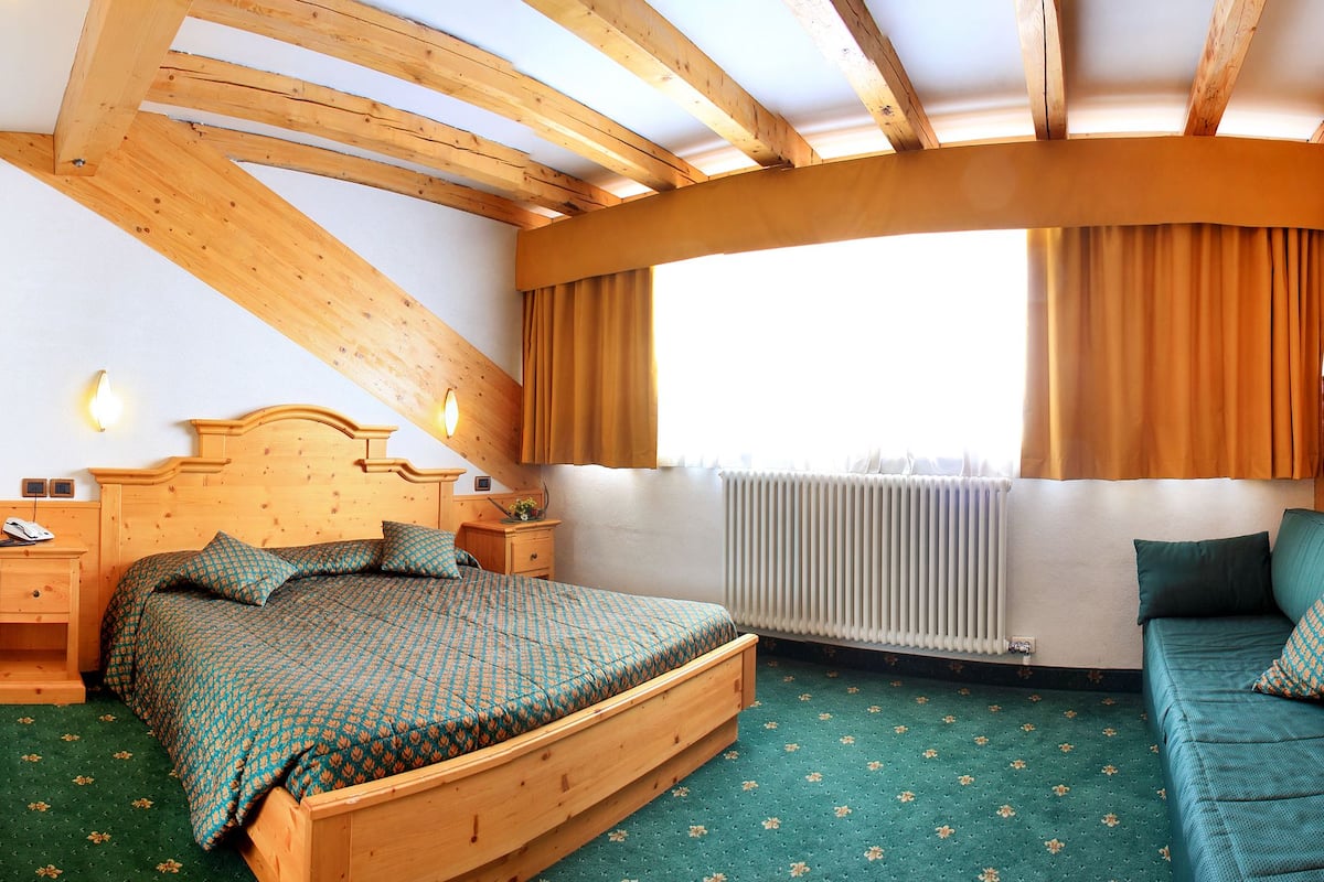 A bedroom with a wooden ceiling, a bed with a patterned bedspread, a nightstand, a dresser, and a window with curtains.