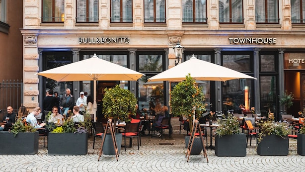 Ein Außenbereich eines Cafés mit Tischen, Stühlen, zwei großen Sonnenschirmen und Blumentöpfen.