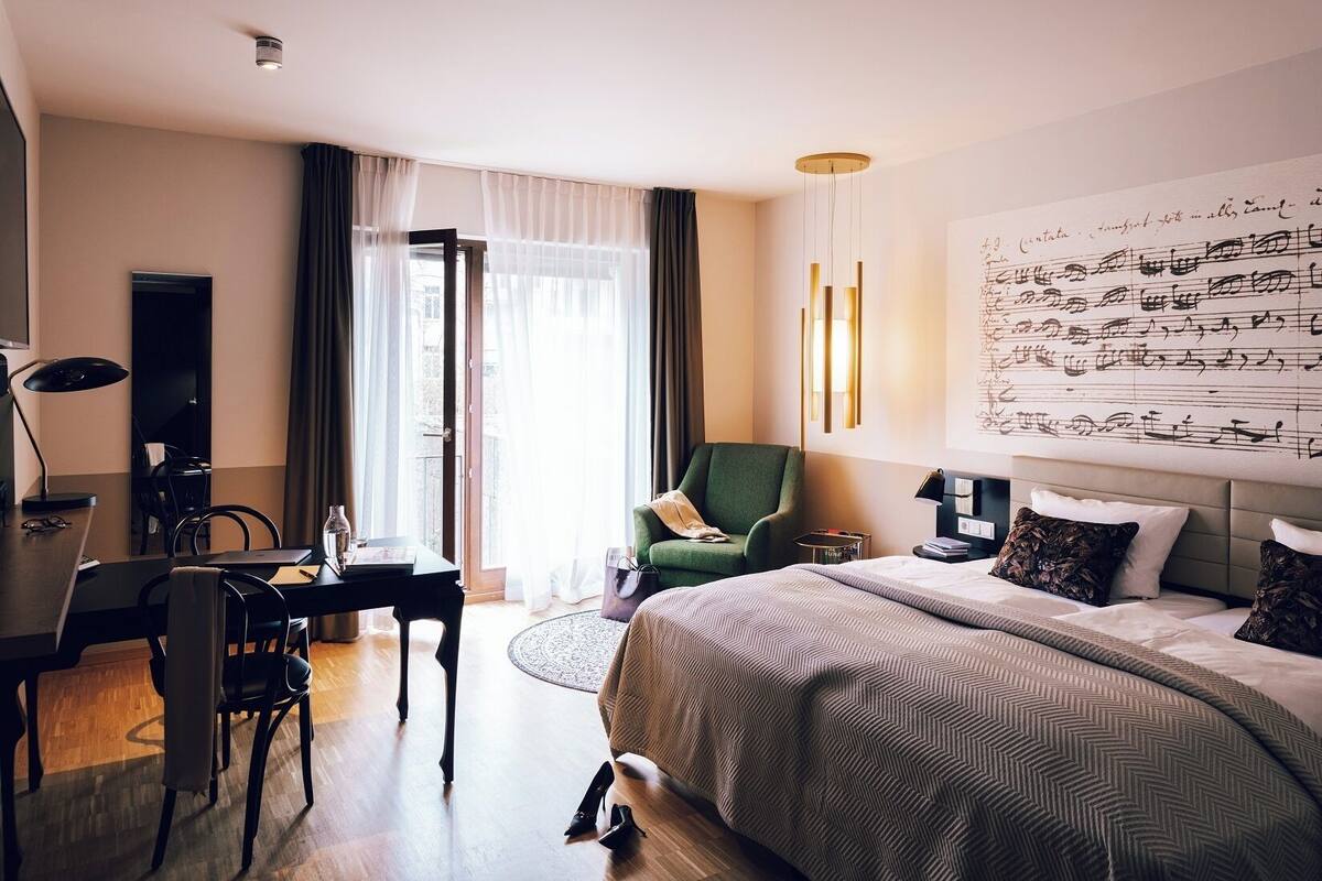 Una habitación de hotel moderna con una cama grande, un escritorio con una silla, un sillón verde y un ventanal con cortinas ligeras.