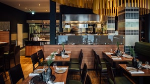 Ein modernes Restaurant mit Holztischen, gepolsterten grünen Sitzbänken und einer sichtbaren Küche.