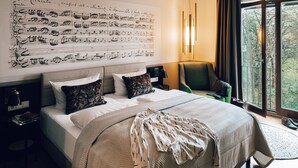 Ein Hotelzimmer mit einem großen Bett, einem Nachttisch, einem Sessel und einem Fenster mit Blick auf Bäume.