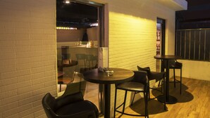 Un espace extérieur aménagé avec des tables rondes et des chaises noires, situé à côté d’un bâtiment dont la façade est en briques et qui possède une fenêtre.
