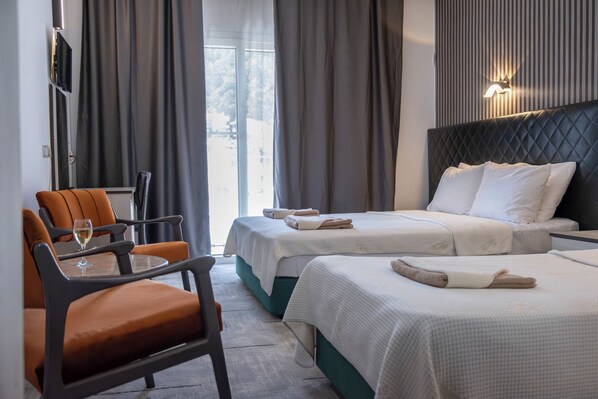Habitación de hotel con dos camas, una zona de estar con una mesa y sillas, y vistas a través del ventanal.