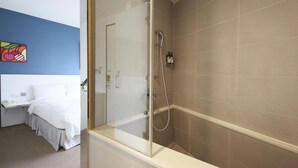 A modern bathroom with a glass-enclosed shower, beige tiles, and a bed with white bedding in the adjacent room.