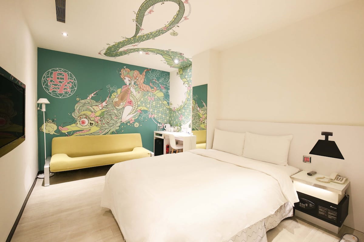 A hotel room with a large bed, a yellow sofa, a desk with a lamp, and a wall mural.