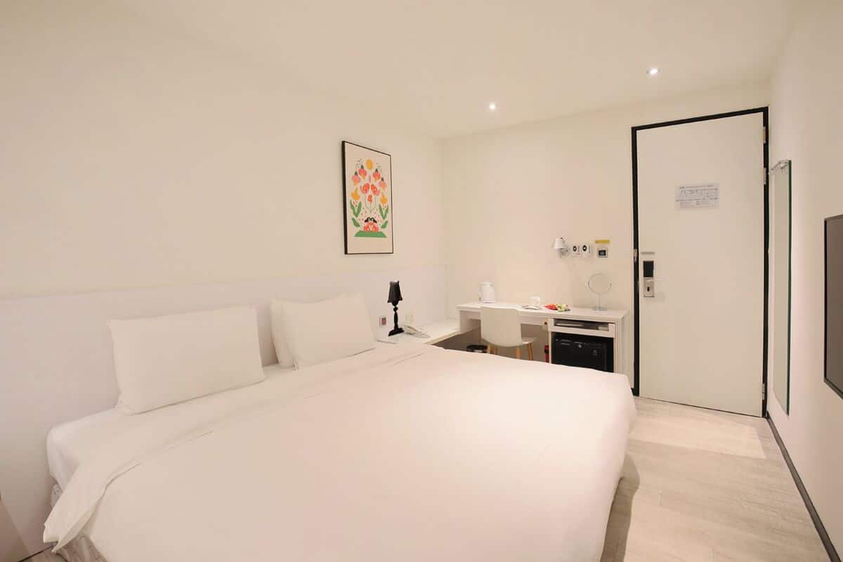 A hotel room with a large bed, a desk, a chair, a desk lamp, a coffee maker, and a door with a sign.