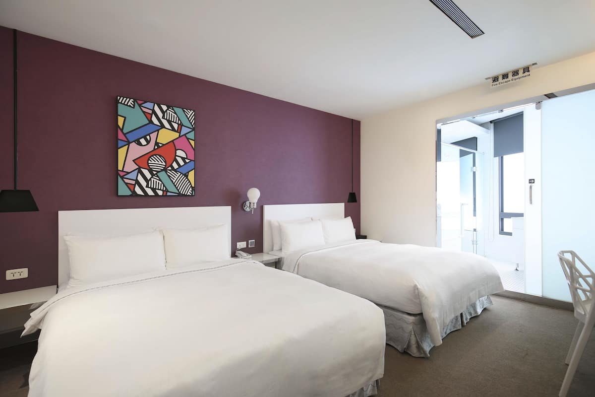A hotel room with two beds, a colorful abstract painting on the wall, a bedside table with a lamp, and a view of the outside through a door.