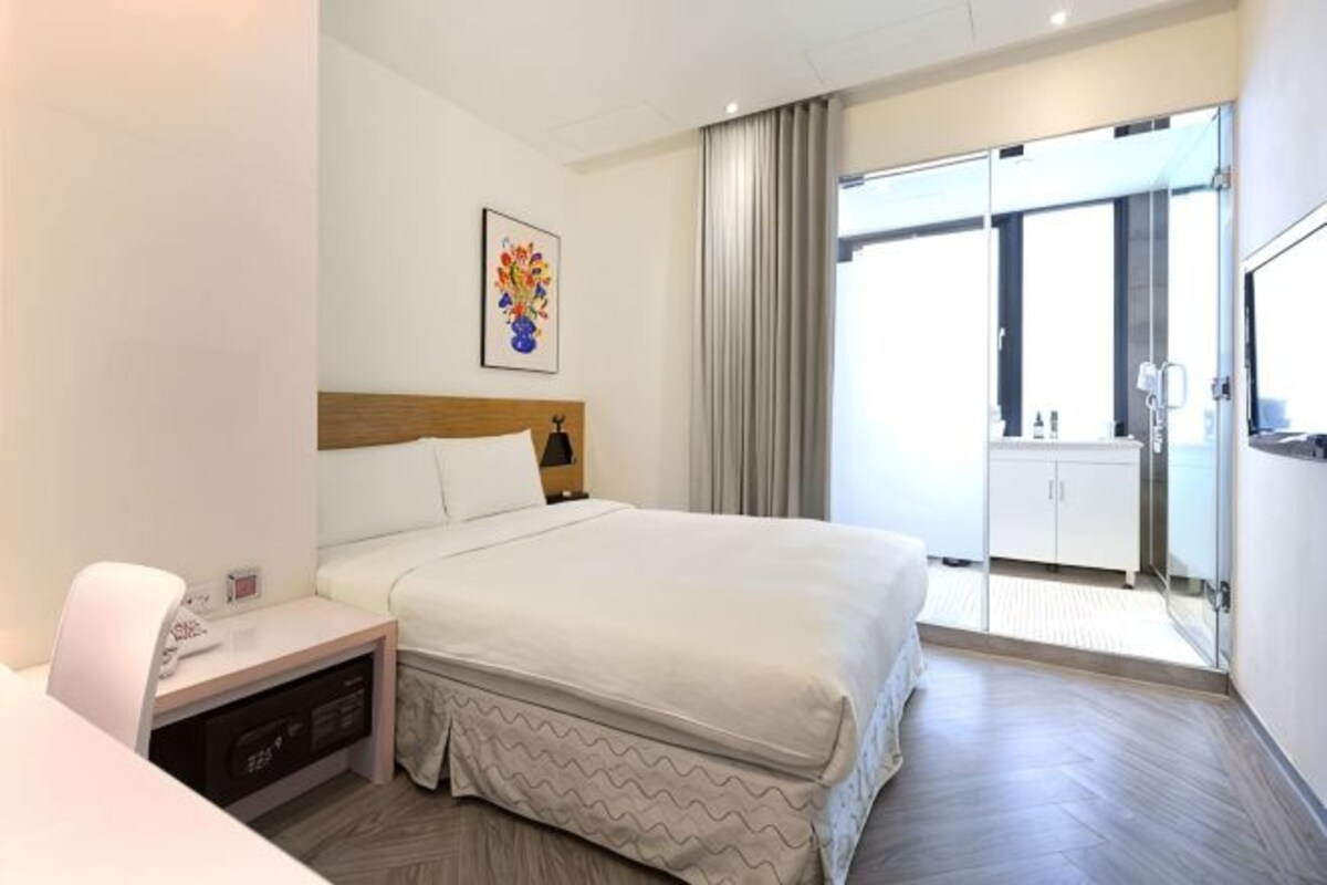 A hotel room with a bed, bedside table, lamp, and a view of the outside.