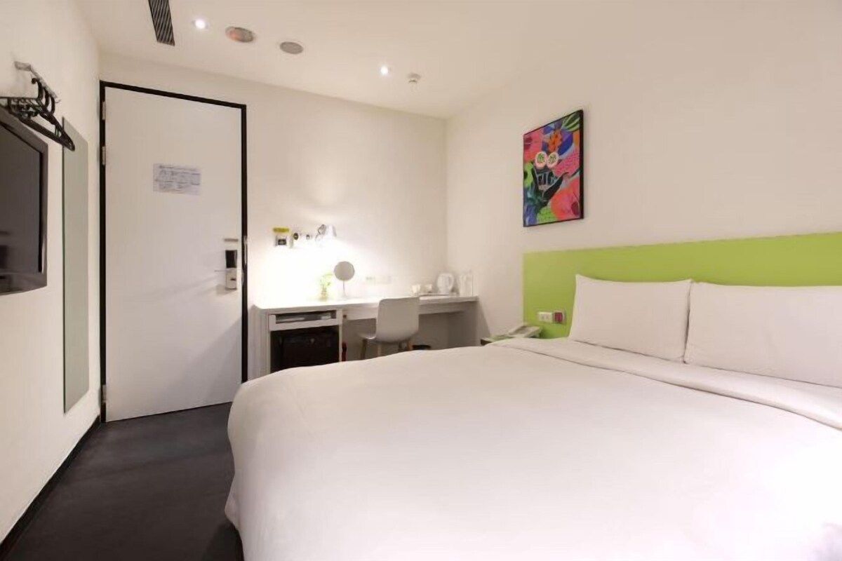 A hotel room with a bed, a desk with a chair, a mirror, and a colorful painting on the wall.