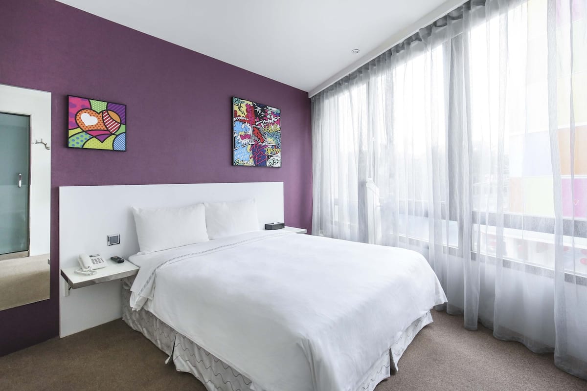 A hotel room with a large bed, a bedside table with a phone, and two colorful artworks on the wall.