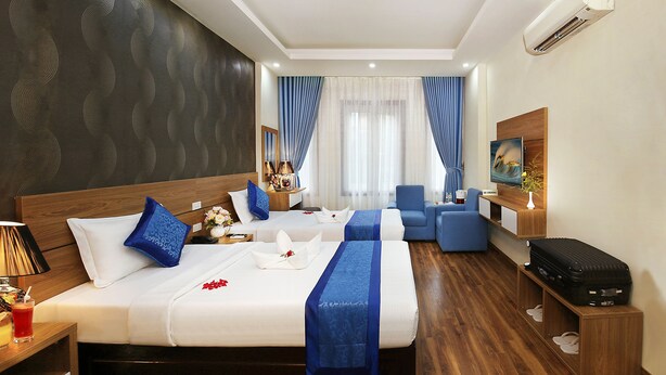 A hotel room with a large bed, a desk, a chair, a TV, and a suitcase on a stand. - Aquarius Grand Hotel (Hanoi)