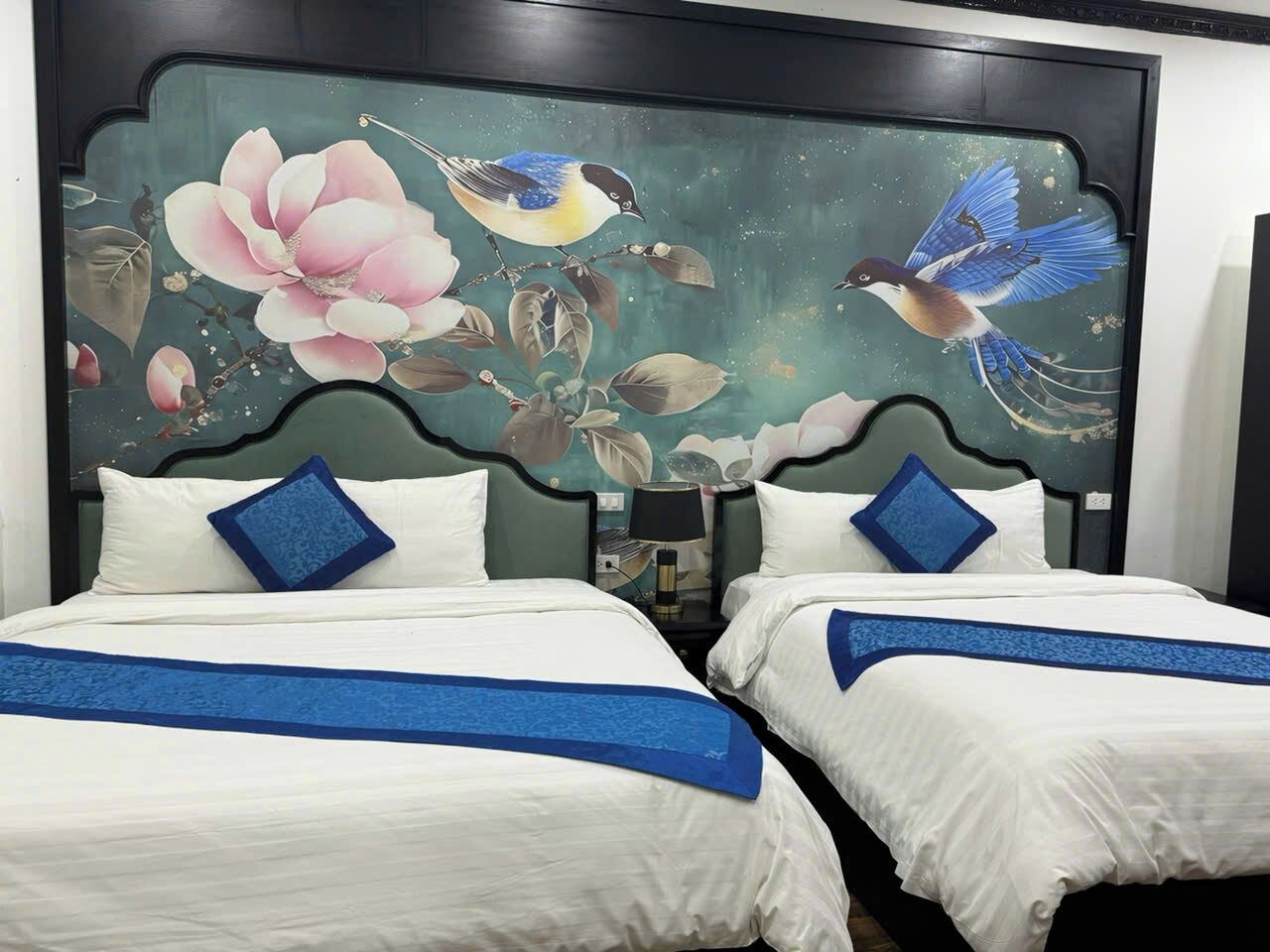 Une chambre d’hôtel avec deux lits, chacun agrémenté de draps blancs et de touches de couleur bleue. Une grande fresque représentant des oiseaux et des fleurs orne la tête de lit.