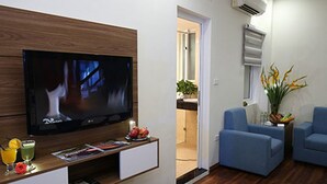 A living room with a flat-screen TV, a coffee table with drinks and magazines, two blue armchairs, and a potted plant. - Aquarius Grand Hotel (Hanoi)