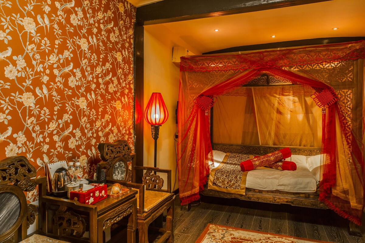 A traditional room with a bed under a red canopy, a wooden desk with a chair, and a wall with floral wallpaper.