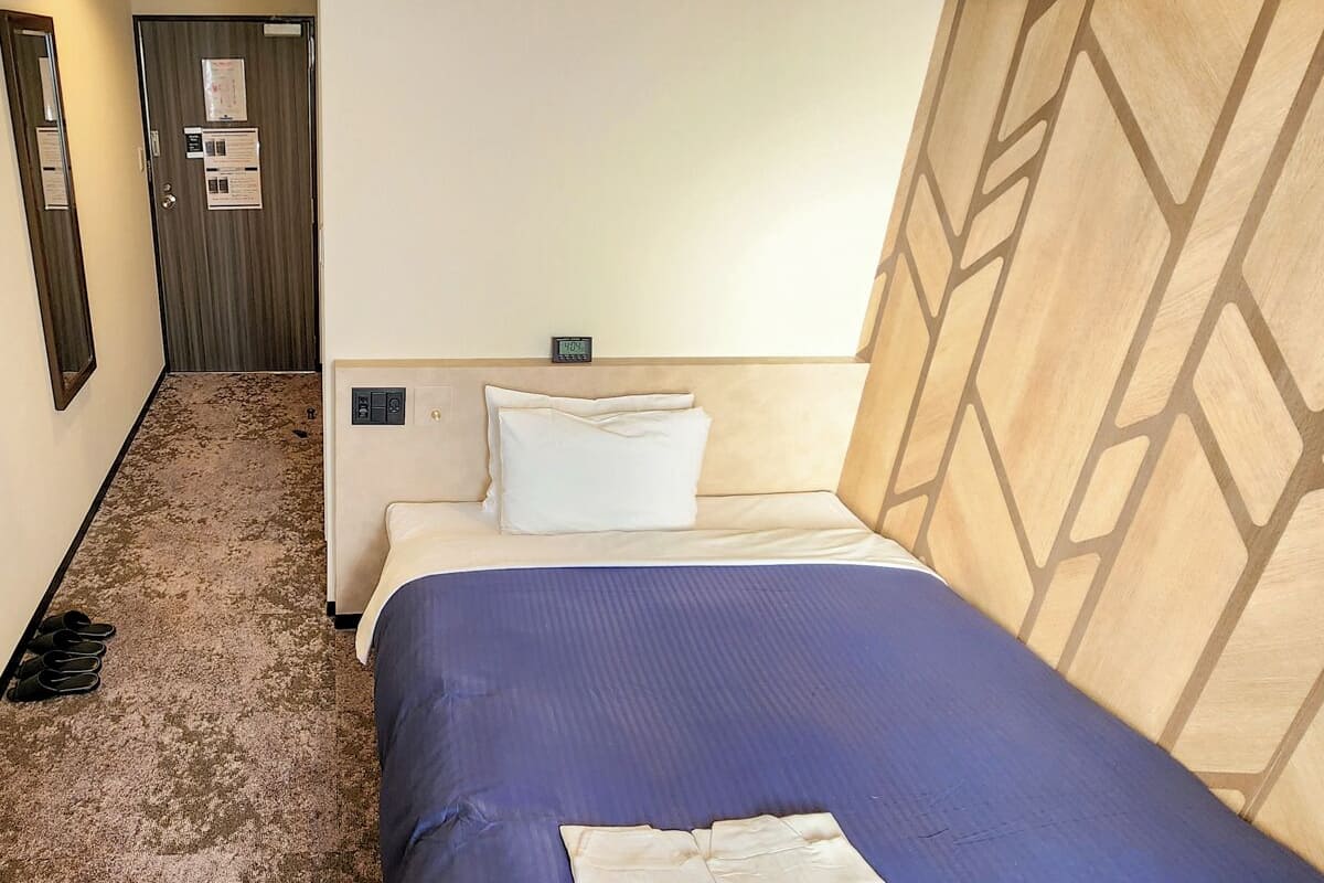 Una habitación de hotel con cama, mesita de noche, reloj y puerta con un aviso.