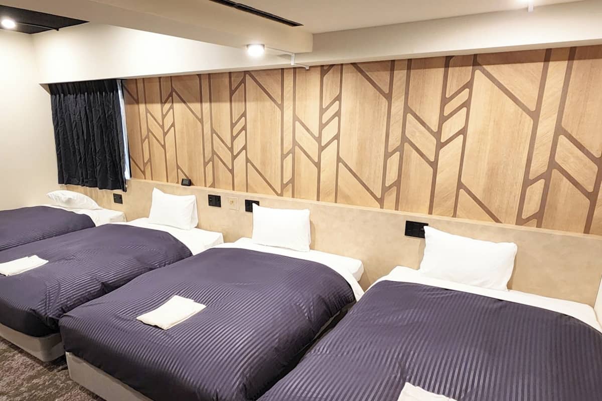 Habitación de hotel con cuatro camas dispuestas en fila, cabecera de madera con diseño geométrico, cortina negra y un pequeño escritorio con teléfono.