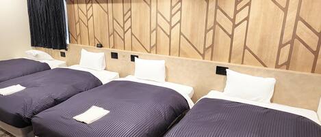 Habitación de hotel con cuatro camas dispuestas en fila, cabecera de madera con diseño geométrico, cortina negra y un pequeño escritorio con teléfono.
