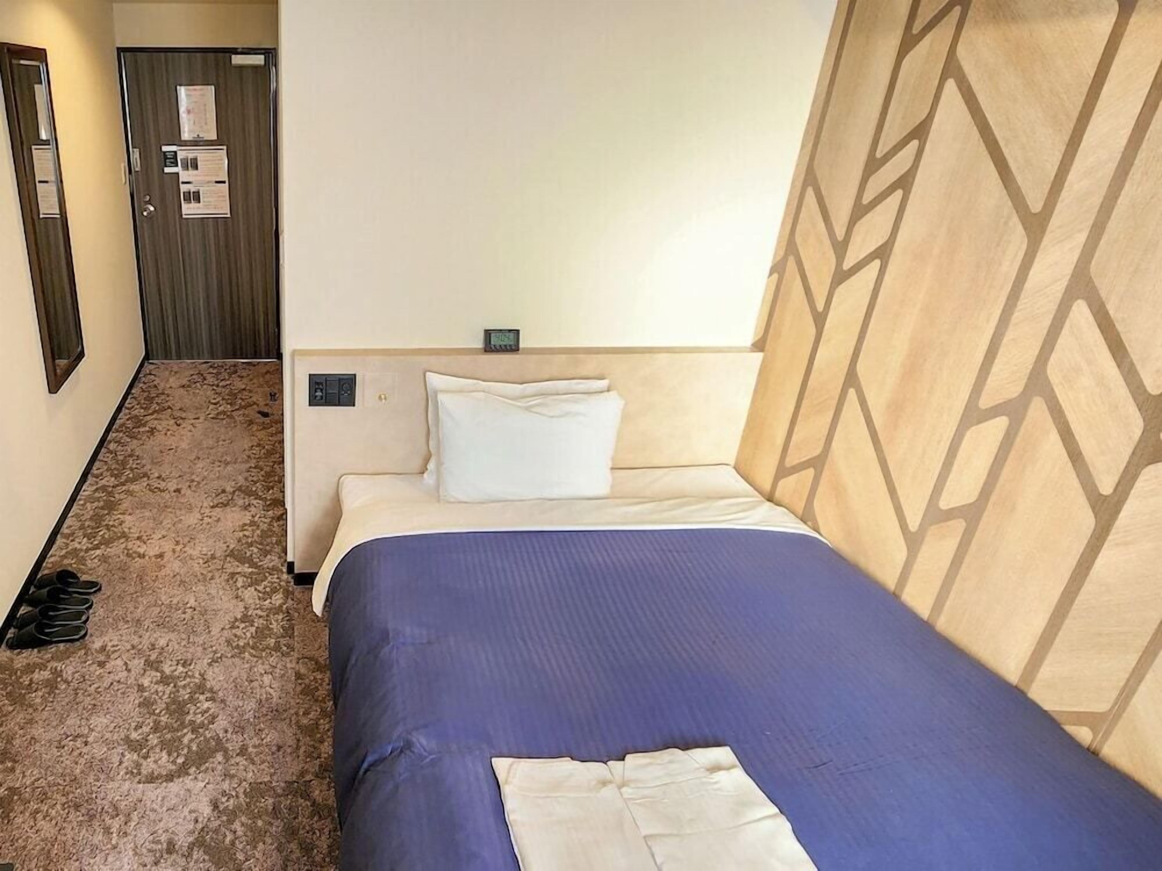 A hotel room with a bed, a headboard with a geometric pattern, a wall clock, and a door with a mirror and a notice.