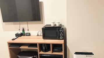 Quarto de hotel compacto com escrivaninha de madeira, micro-ondas, lixeira pequena e TV fixada na parede.