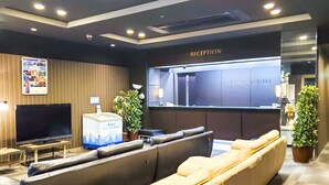 A modern reception area with a reception desk, a flat-screen TV, a seating area with a sofa and armchairs, and a large potted plant. - HOTEL LiVEMAX Hiratsuka Ekimae (Hiratsuka)