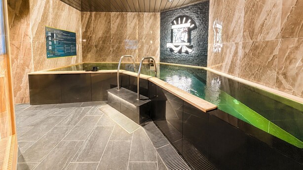 A modern indoor swimming pool with a stone wall, a wooden ceiling, and a sign with text. - HOTEL LiVEMAX Hiratsuka Ekimae (Hiratsuka)