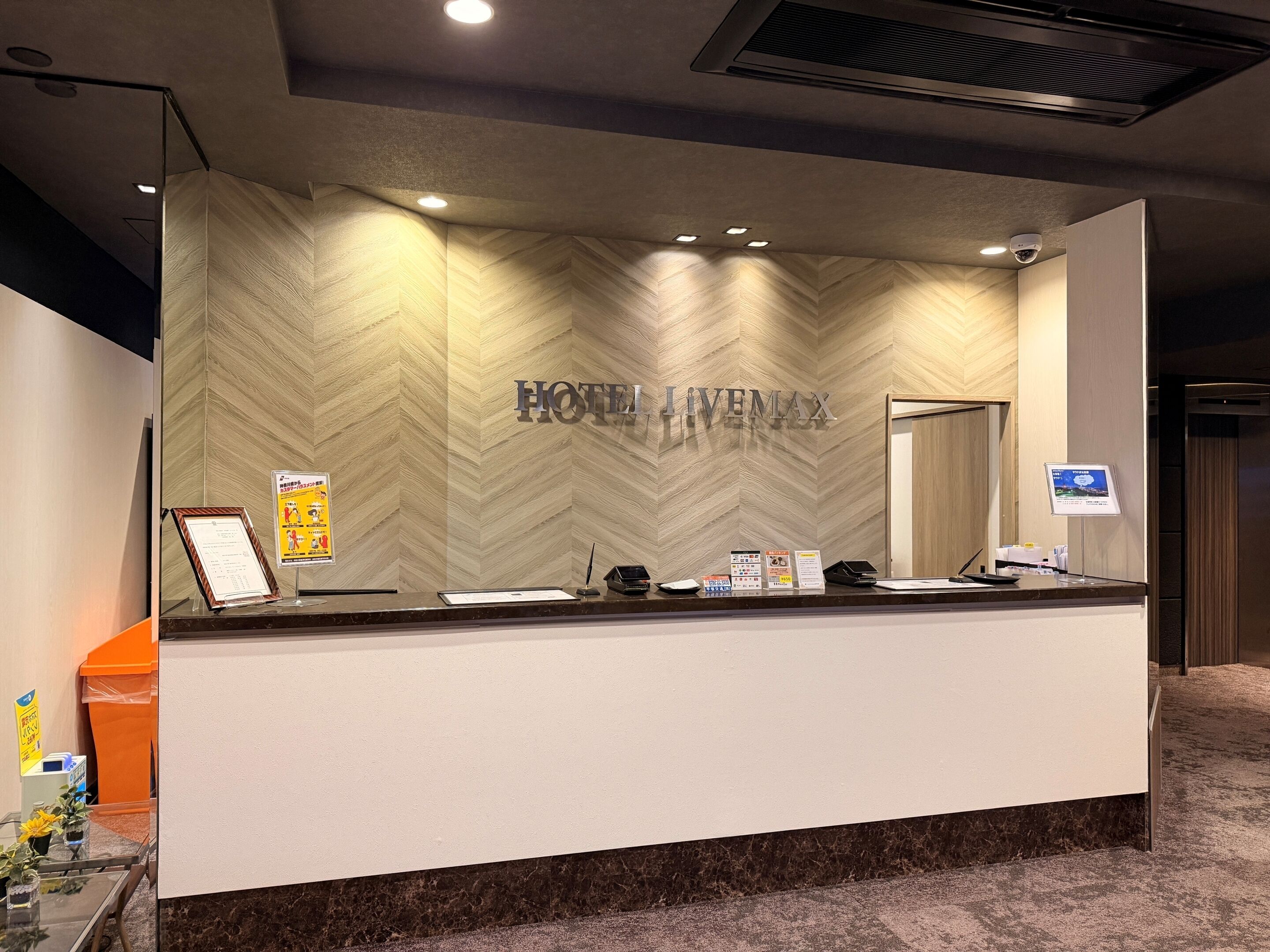 A hotel reception desk with a herringbone-patterned wall, a sign reading 'HOTEL LIVEMAX,' and various informational posters.