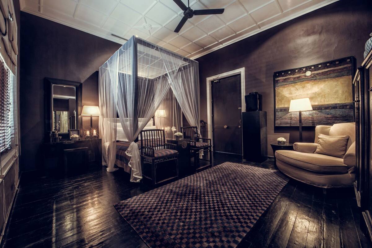 Une chambre à coucher avec un lit à baldaquin, un coin salon avec un canapé et des fauteuils, un grand tableau au mur et un ventilateur de plafond.