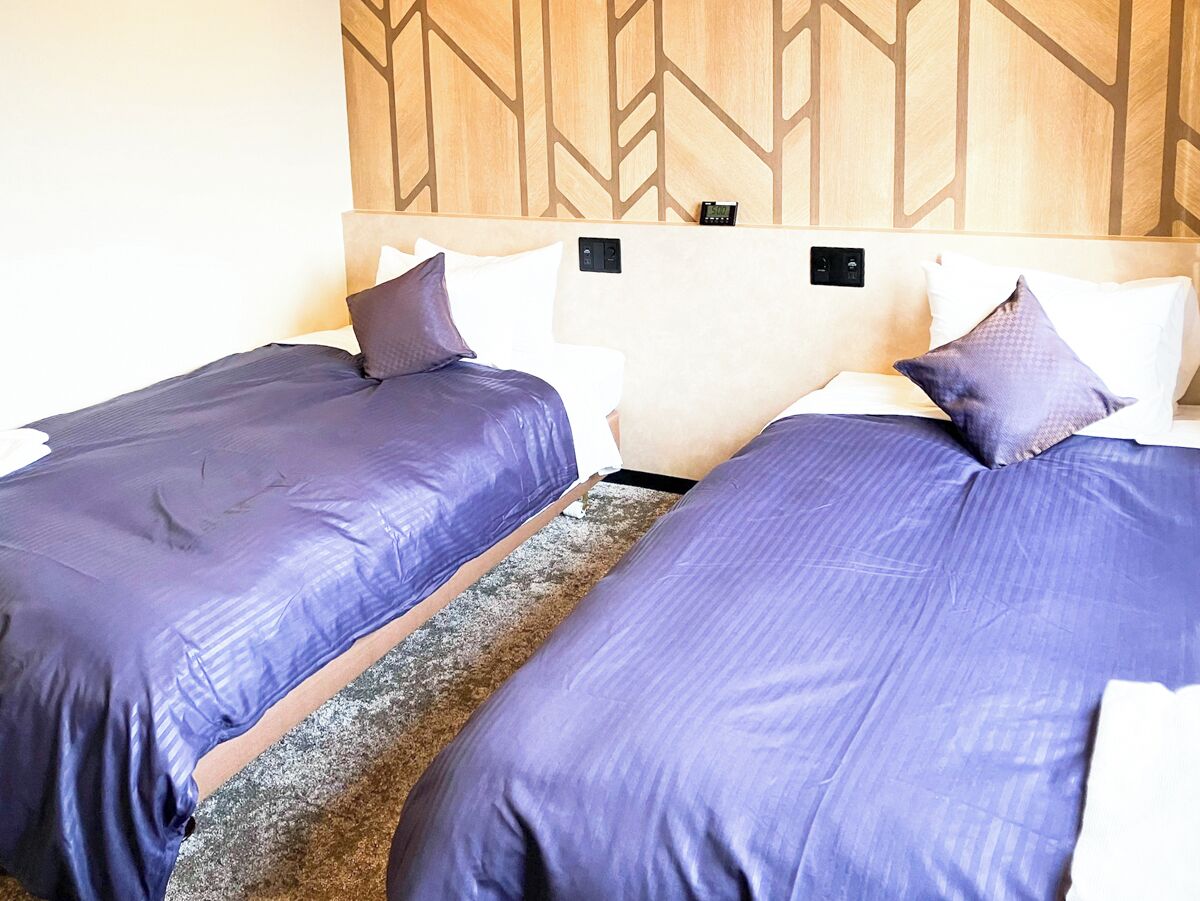 Ein Hotelzimmer mit zwei Betten, jeweils mit lila Bettdecke und passenden Kissen. Das Zimmer verfügt über einen hölzernen Kopfseitenverkleidung mit geometrischem Muster und eine kleine Bedienkonsole über den Betten.