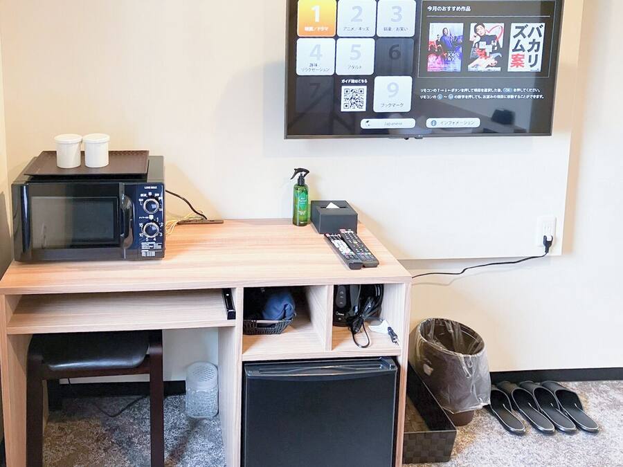 A compact kitchenette with a microwave, two small containers, a remote control, and a trash bin.