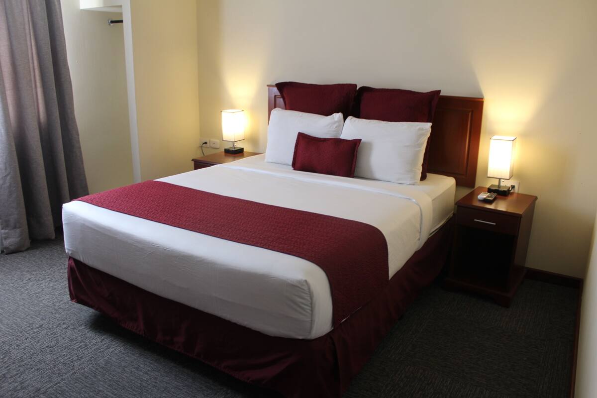 A hotel room with a large bed, two bedside tables with lamps, a wooden headboard, and a carpeted floor.