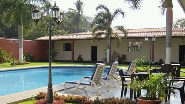 A pool area with lounge chairs and tables, surrounded by palm trees and a building with a tiled roof.