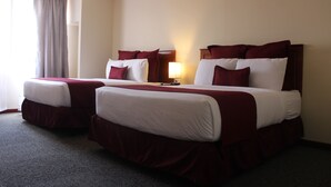 A hotel room with two beds, each with white and red bedding, a bedside table with a lamp, and a large window with curtains.
