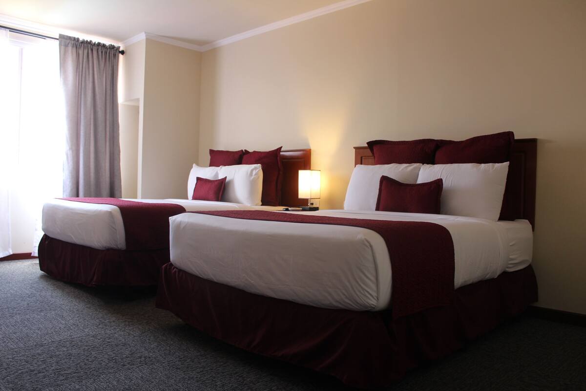 A hotel room with two beds, each with white and red bedding, a bedside table with a lamp, and a large window with curtains.