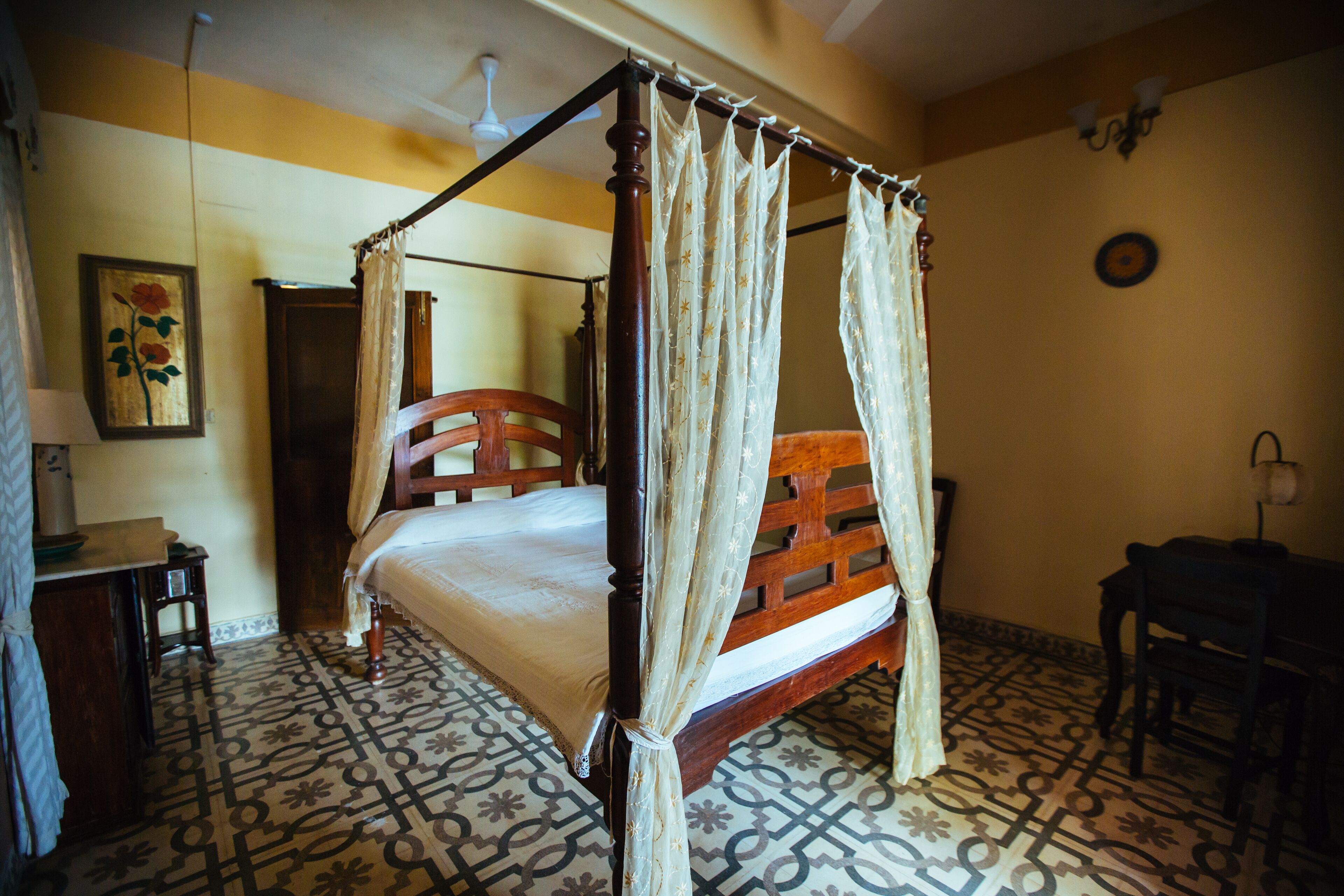 Un lit à baldaquin avec une structure en bois et des rideaux blancs, une chaise en bois, un bureau avec une lampe et des carreaux de sol à motifs.