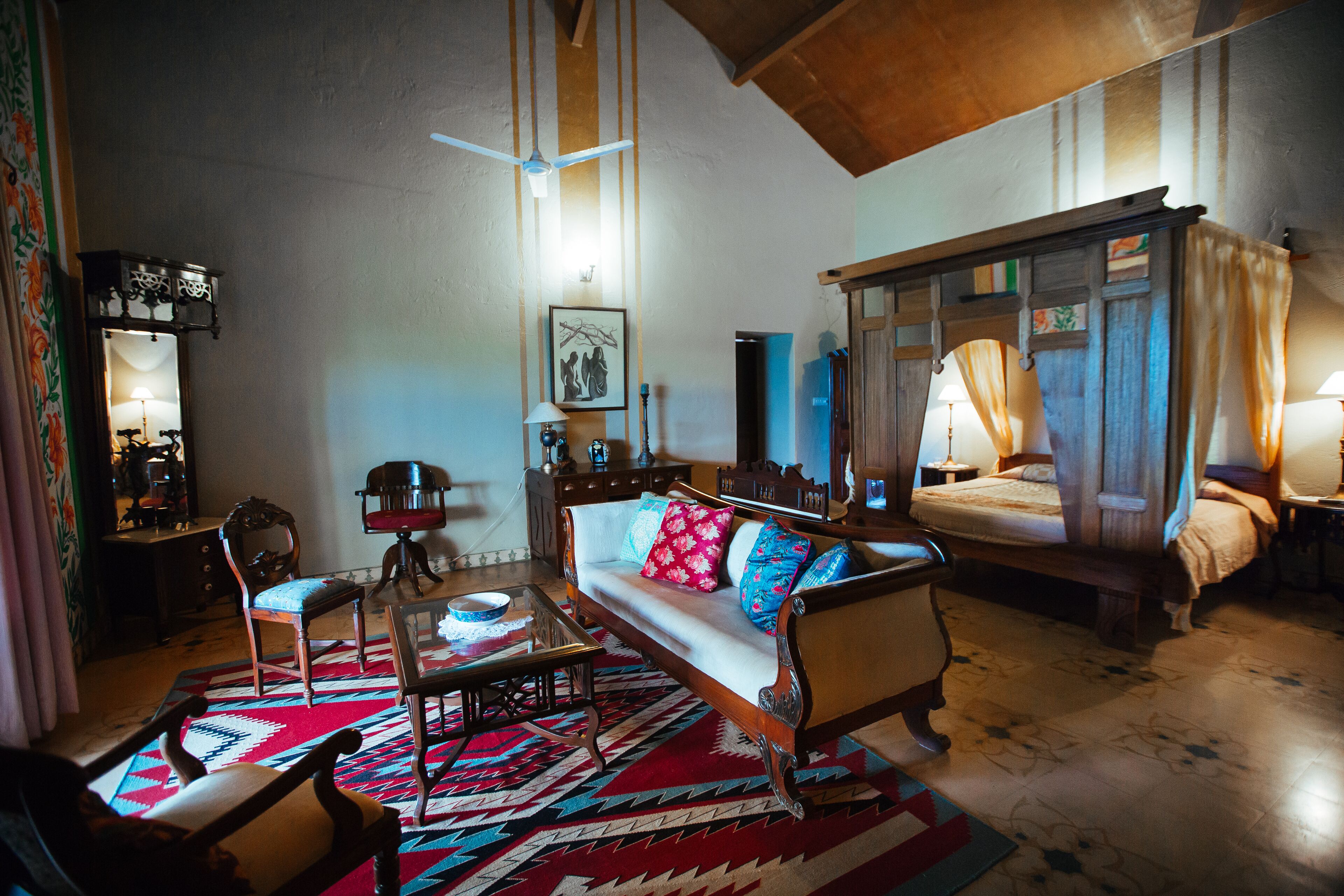 Una habitación tradicional con sofá, una silla de madera, una mesa de centro de vidrio, alfombra estampada y cama con dosel.