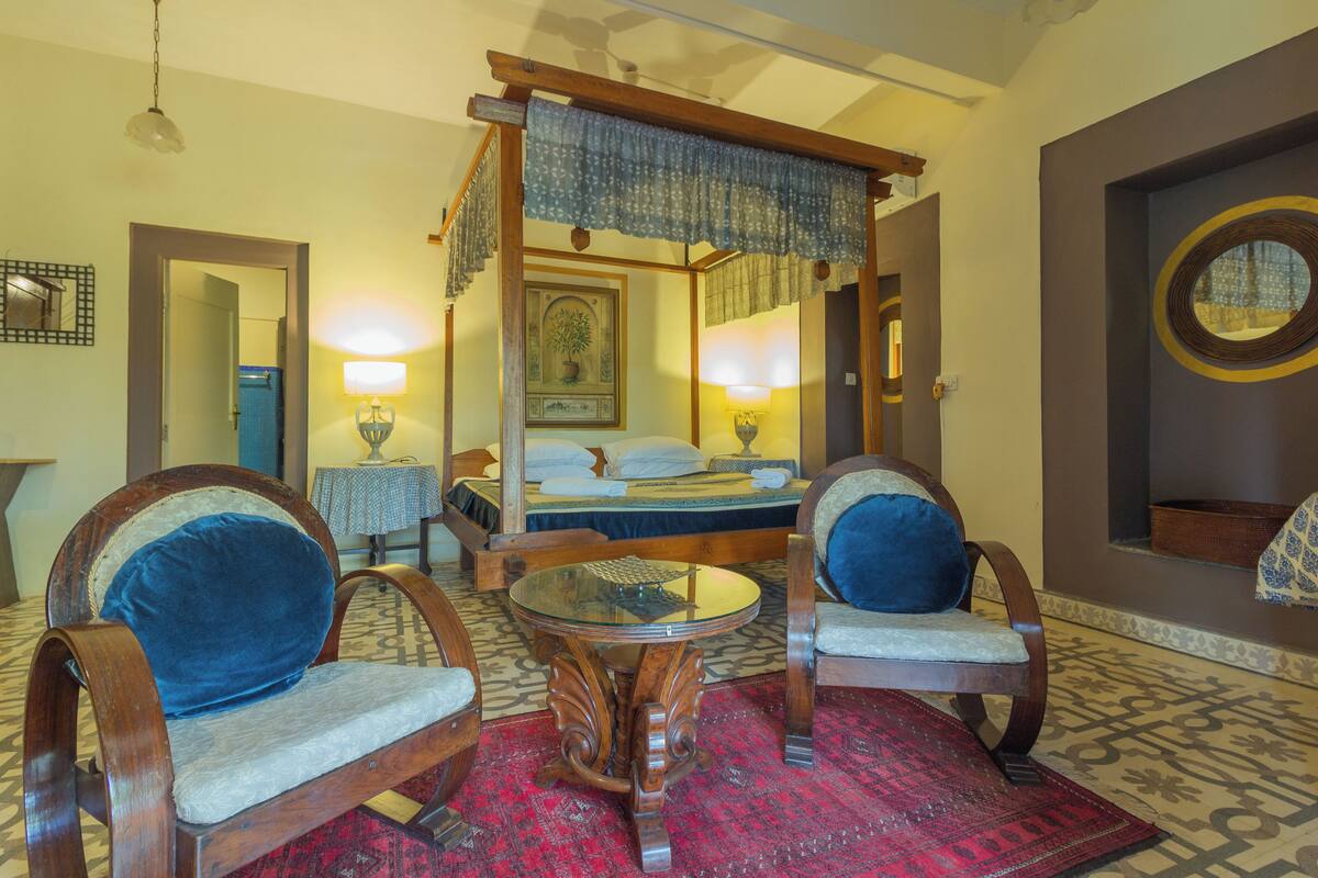 A traditional four-poster bed with a canopy, two upholstered chairs with blue cushions, a round glass table, and a patterned red rug.