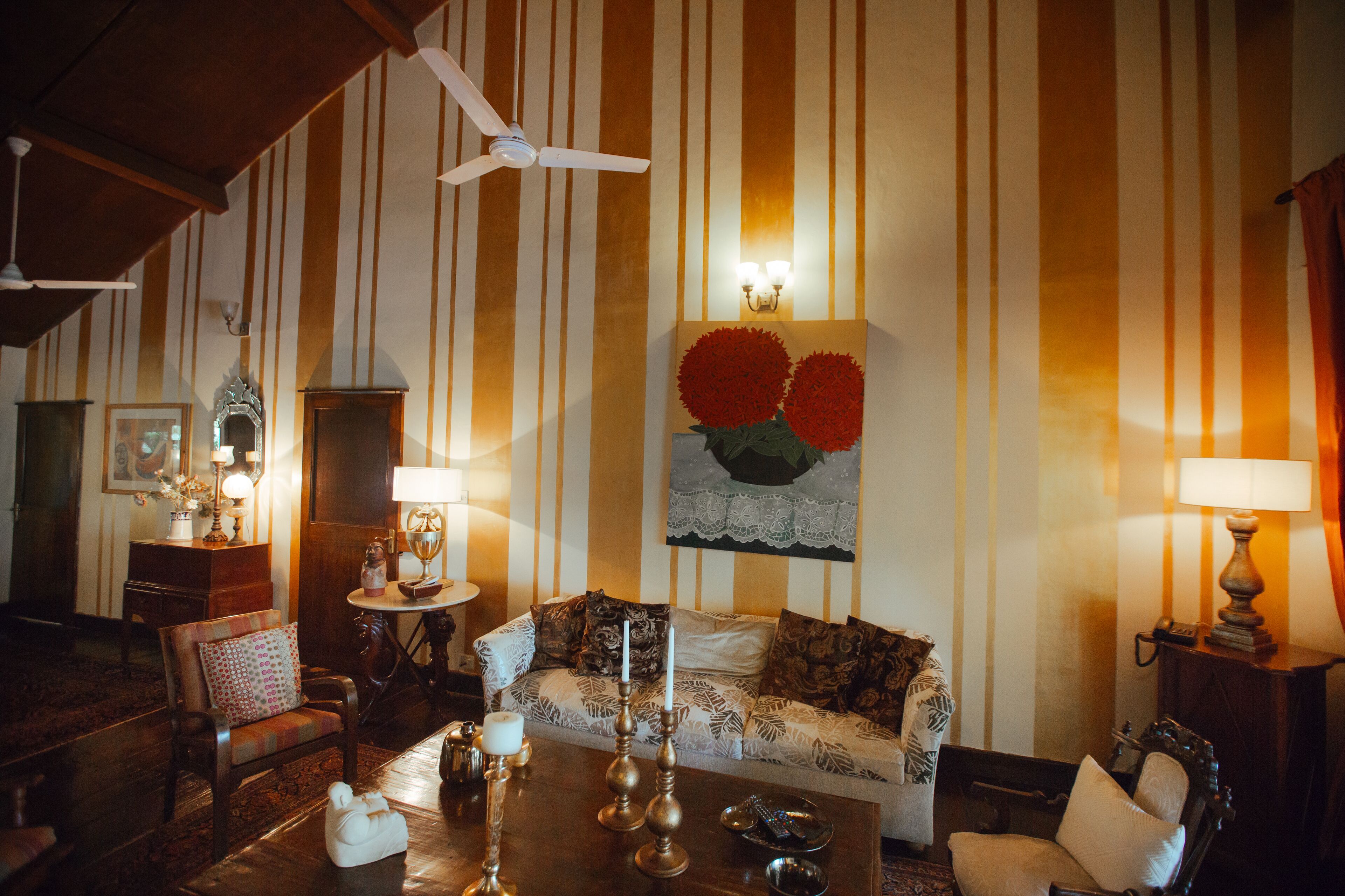 Quarto com papel de parede listrado, ventilador de teto, sofá com almofadas estampadas, mesa de centro com objetos decorativos e luminária fixada na parede.