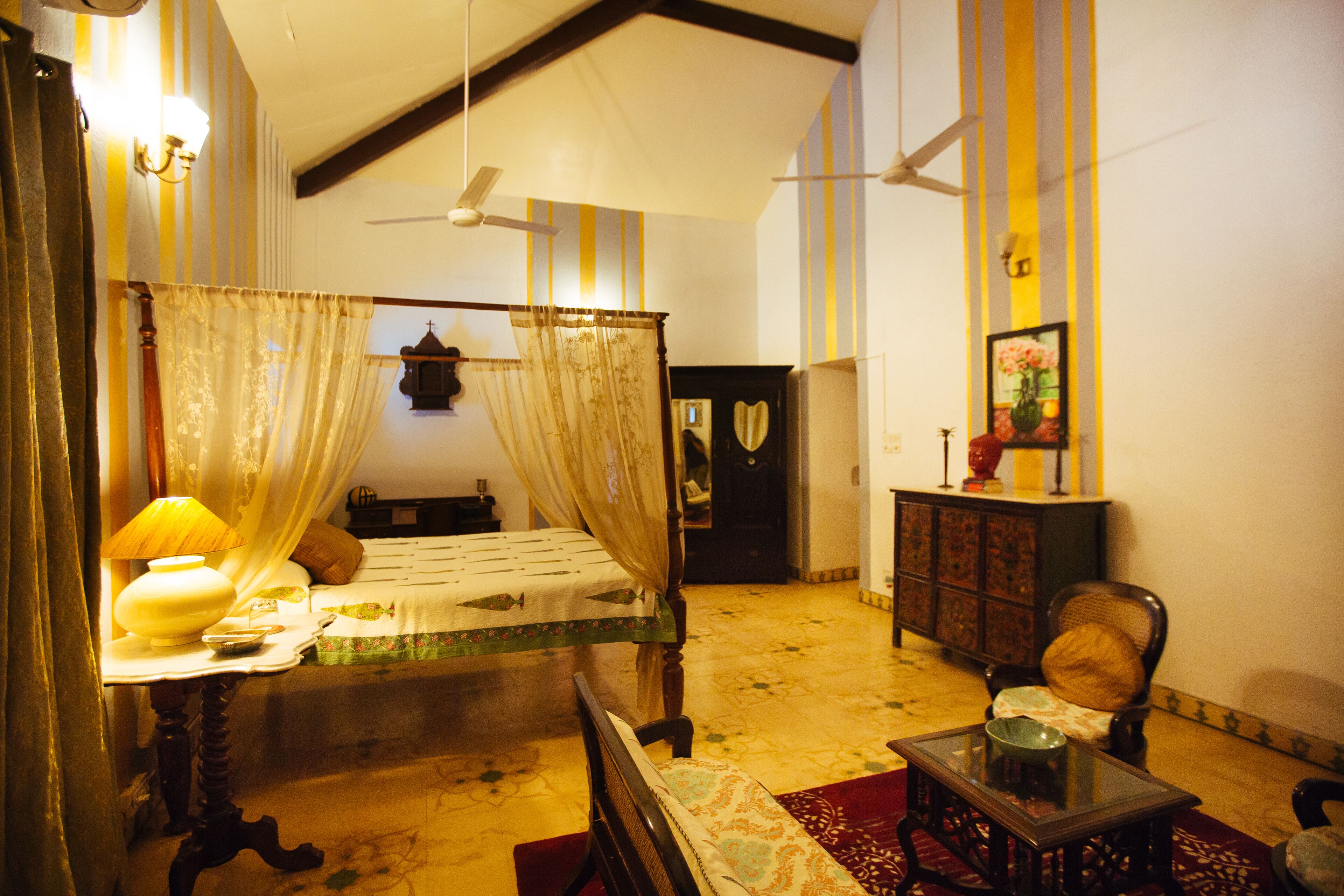 A room with a four-poster bed, a table with a lamp, a chair, and a cabinet.