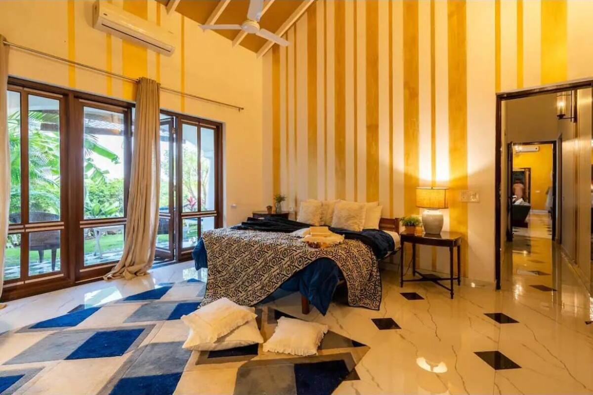 A bedroom with a bed, a nightstand, a lamp, and a view of greenery through large windows.