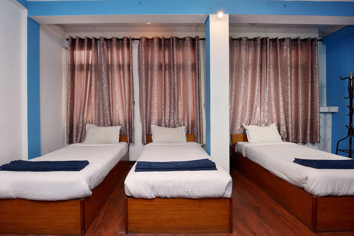 Ein Hotelzimmer mit drei Einzelbetten, jeweils mit einem Holz-Kopfteil, weißer Bettwäsche und einer dunkelblauen Decke. Das Zimmer verfügt über eine blaue Akzentwand und ein großes Fenster mit braunen Vorhängen.