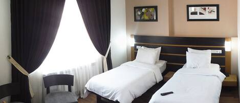A hotel room with two single beds, a small table, a chair, and a window with curtains.