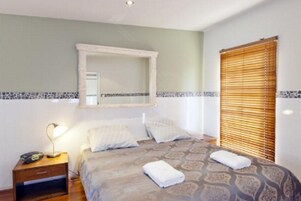 Quarto com cama, mesa de cabeceira, espelho e janela com persianas.