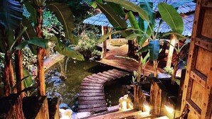 Eine Holzbrücke über einem Teich, umgeben von üppigem Grün und einem Gebäude mit einem Reetdach.