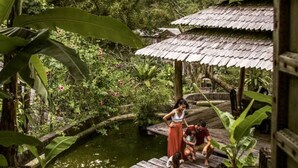 Eine Holzbrücke über einem Teich, umgeben von üppigem Grün und einem traditionellen Gebäude mit einem Reetdach.
