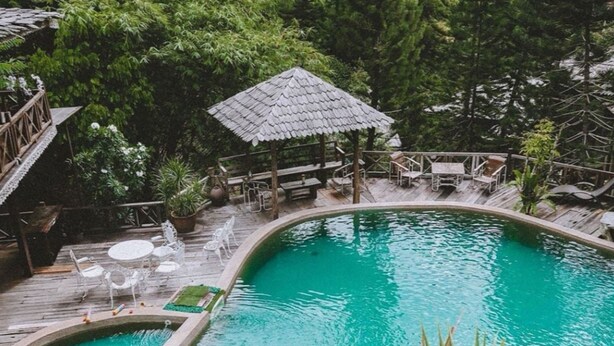 Ein Pool, umgeben von üppiger Vegetation, eine Holzterrasse mit Stühlen und ein Pavillon mit Strohdach.
