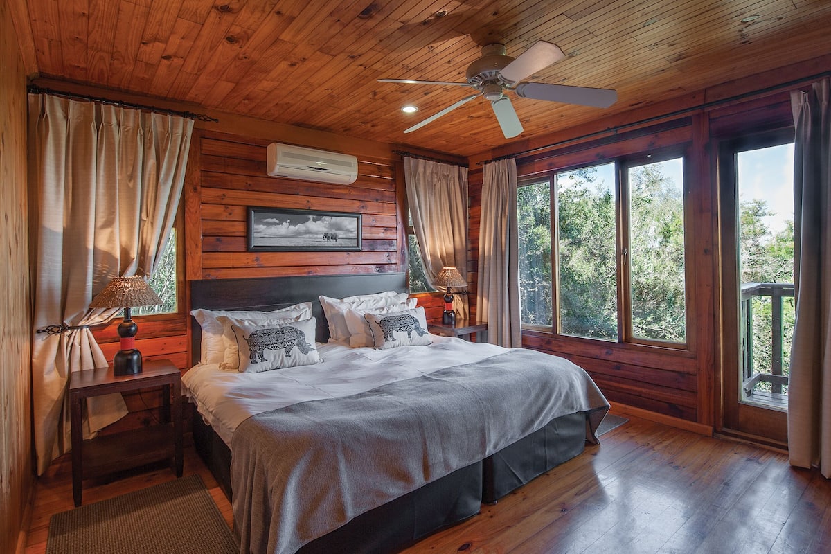 Une chambre dans une cabane en bois, avec un lit, des tables de chevet, un ventilateur de plafond et de grandes fenêtres.